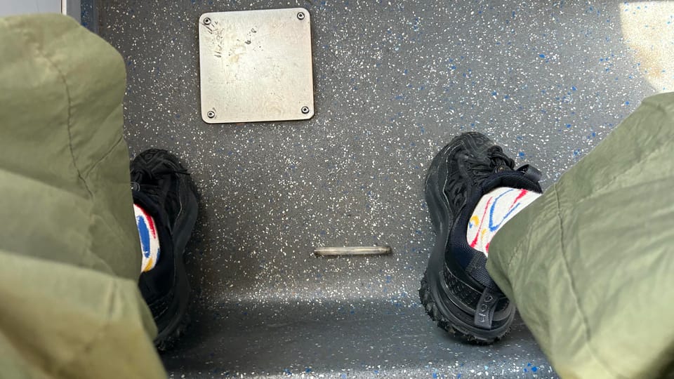Small glass pipe with burnt ends (makeshift crack pipe) laying on grey bus floor, in between writer's feet (wearing black rubber shoes)